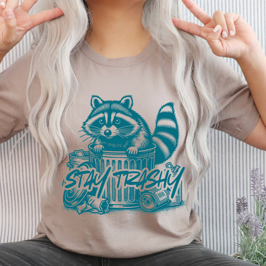 Taupe shirt that has a raccoon on the front in a trash can and the saying stay trashy