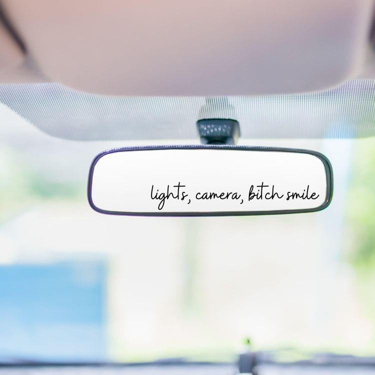 A car decal on the rear view mirror that says lights, camera, bitch smile.