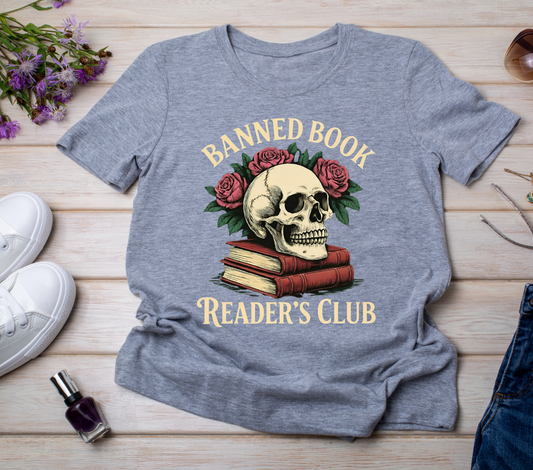 Gray t-shirt with skull and books design on a wooden surface with shoes and flowers.