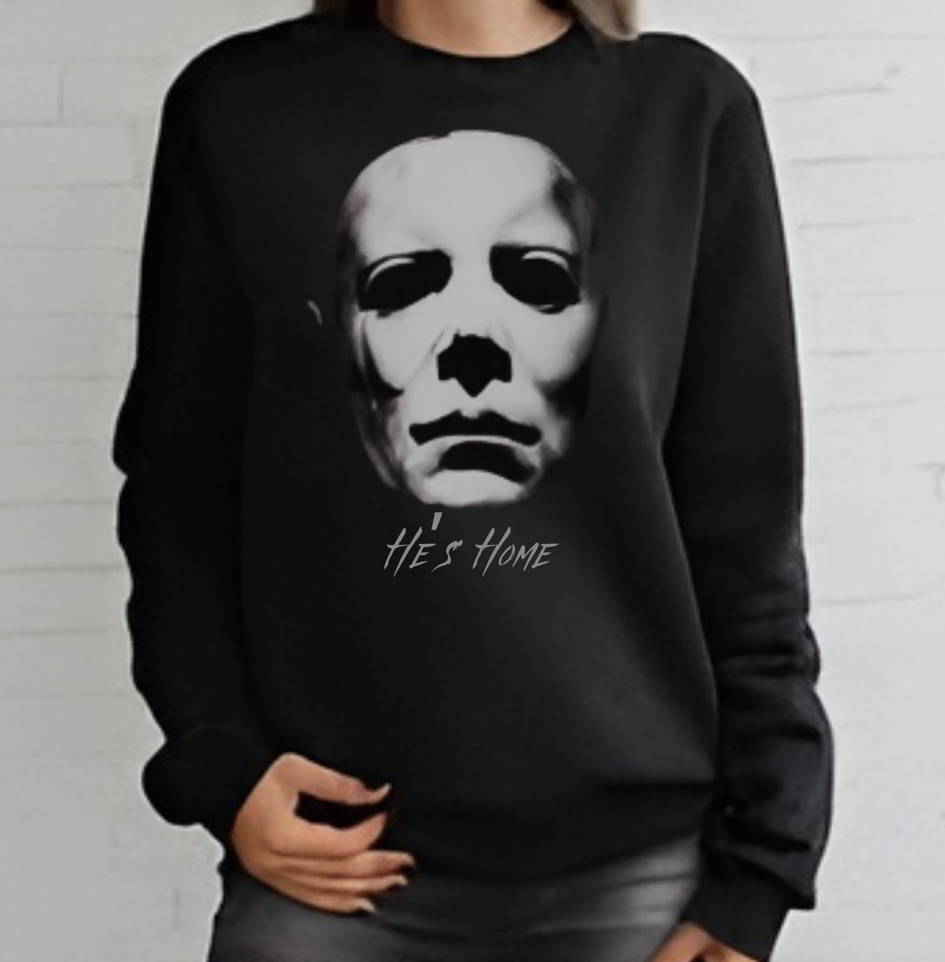 Black Sweatshirt featuring michael myers mask fading into the background ominously and the words, "He's Home" underneath 