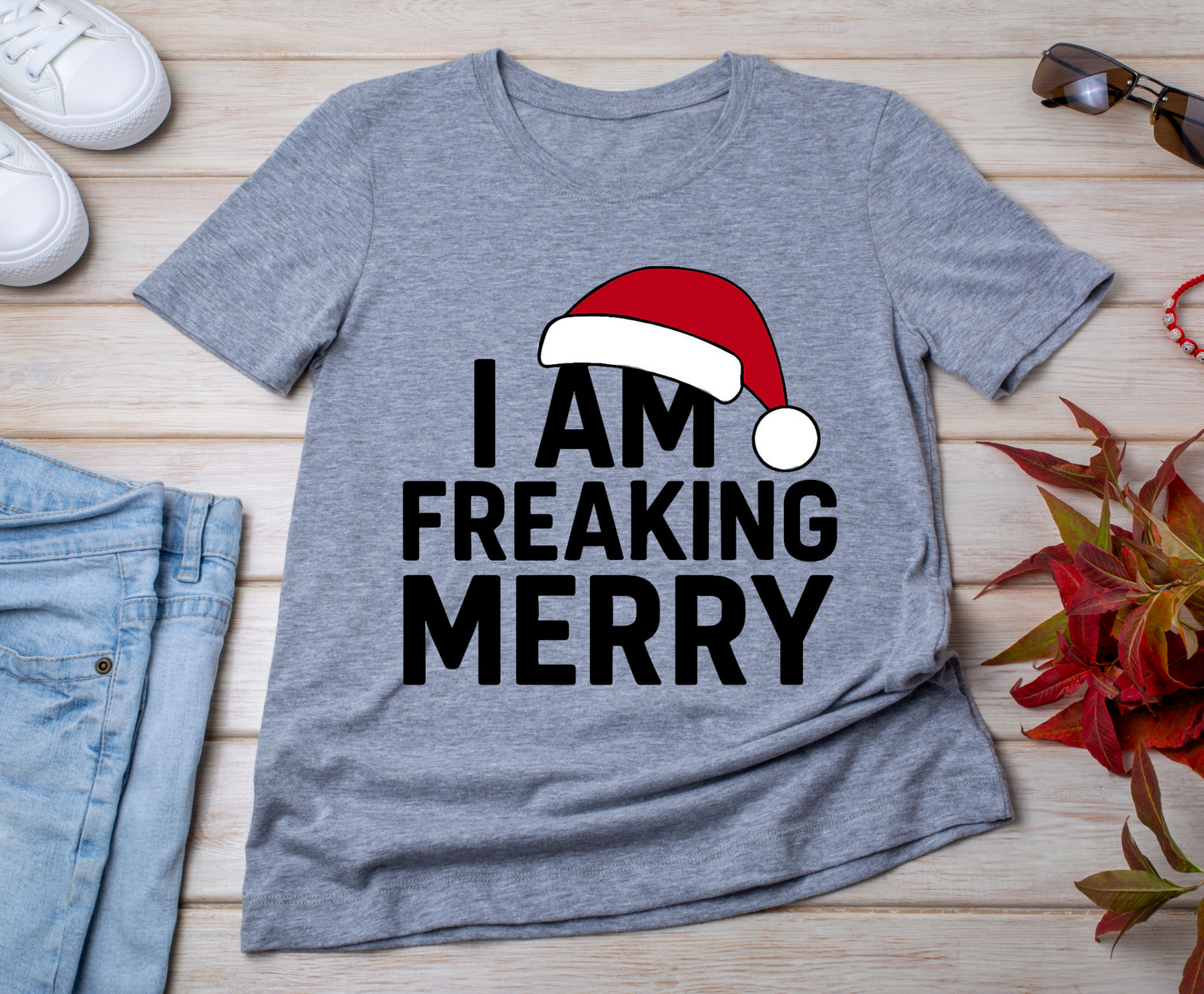 Gray t-shirt with 'I AM FREAKING MERRY' text and Santa hat graphic on a wooden surface with jeans and shoes.