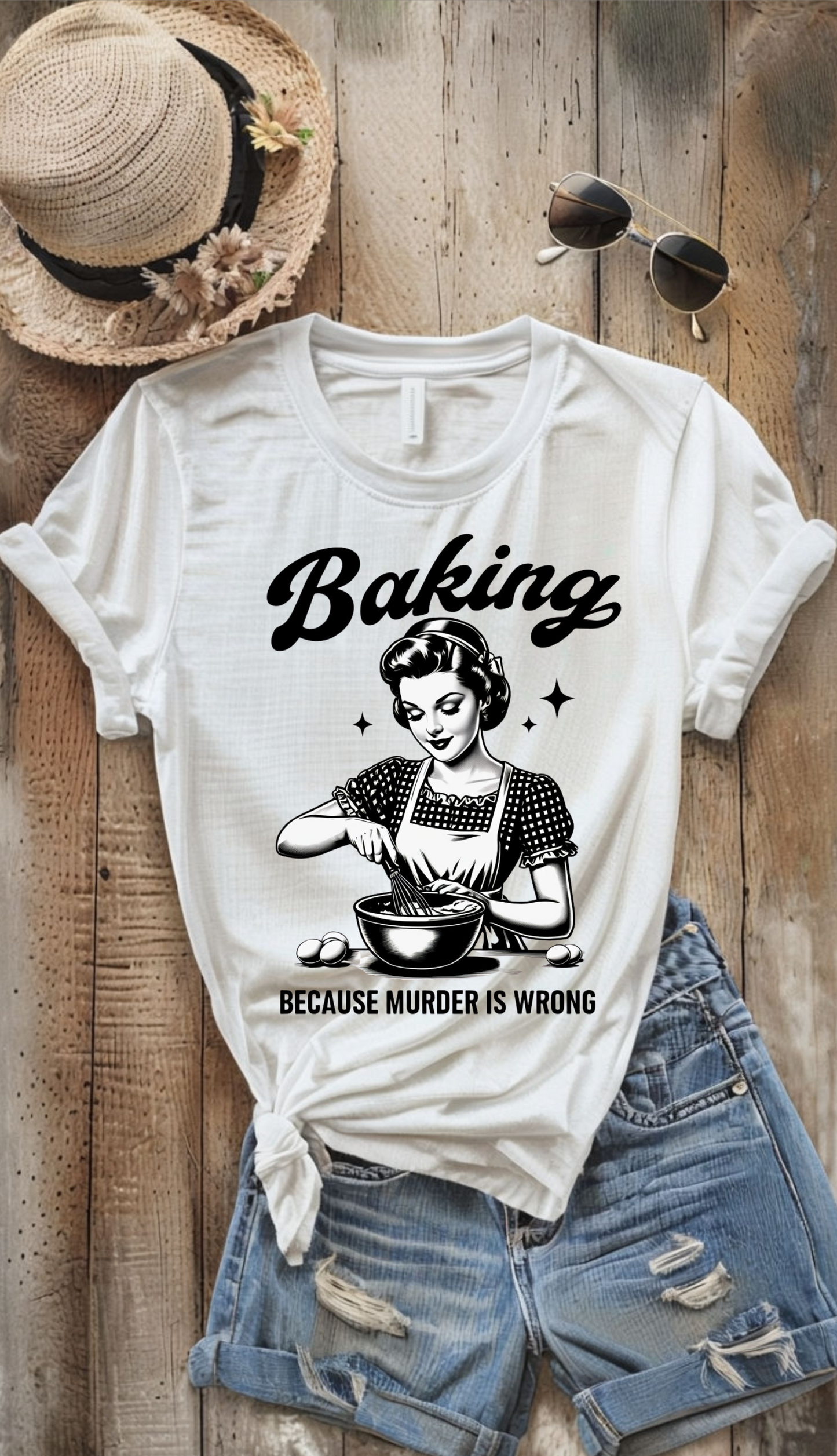 White t-shirt with 'Baking because murder is wrong' graphic on a wooden surface with a hat and sunglasses.