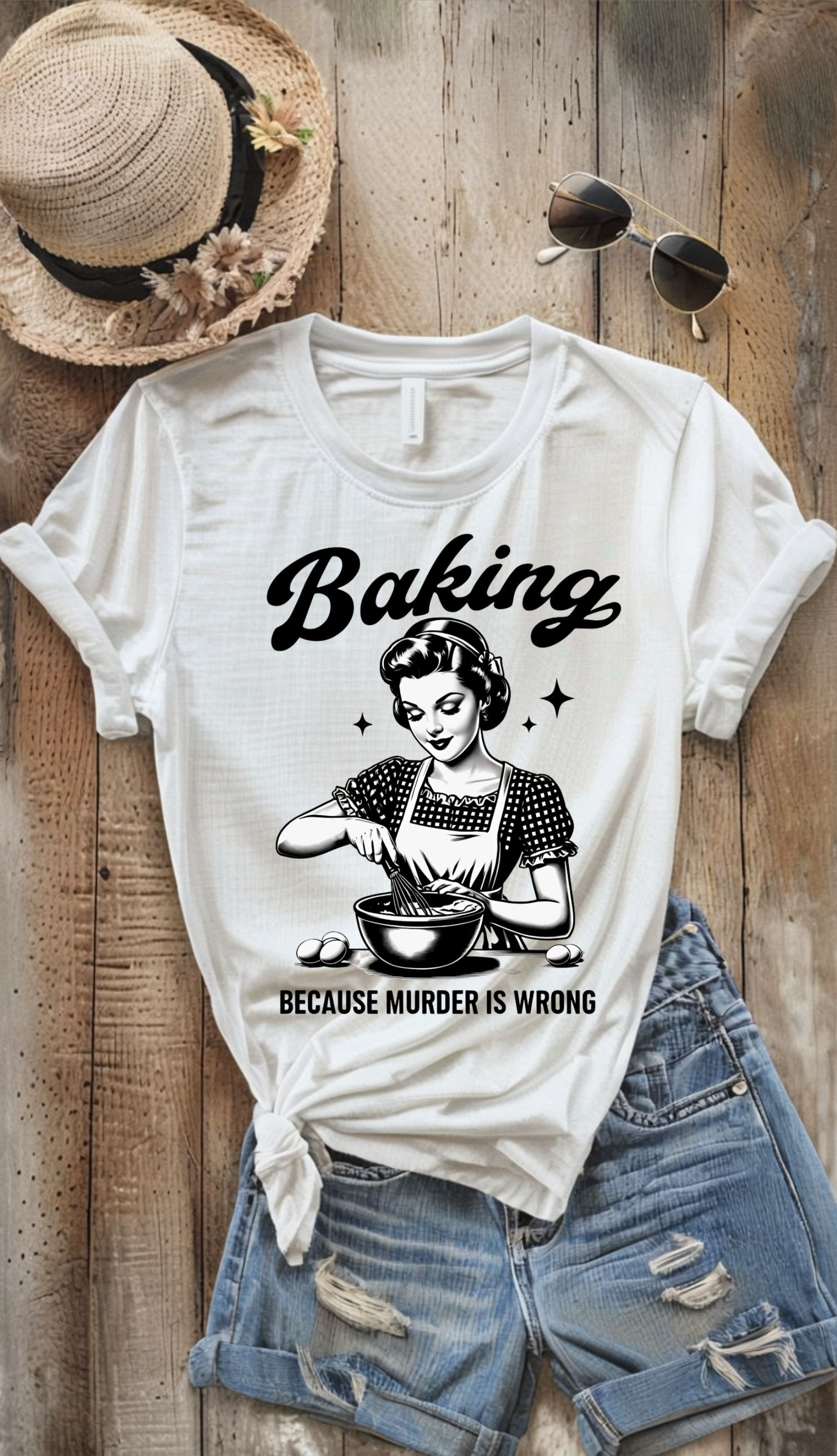 White t-shirt with 'Baking because murder is wrong' graphic on a wooden surface with a hat and sunglasses.
