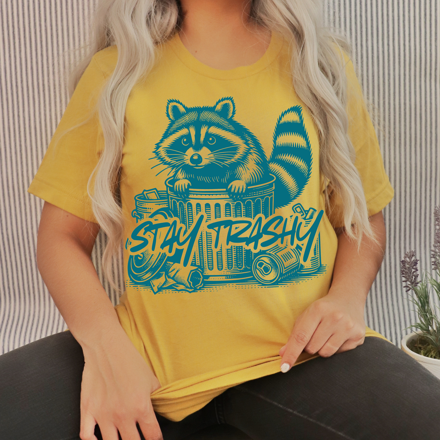 Yellow shirt with a raccoon on the front in an aqua green color and the saying stay trashy