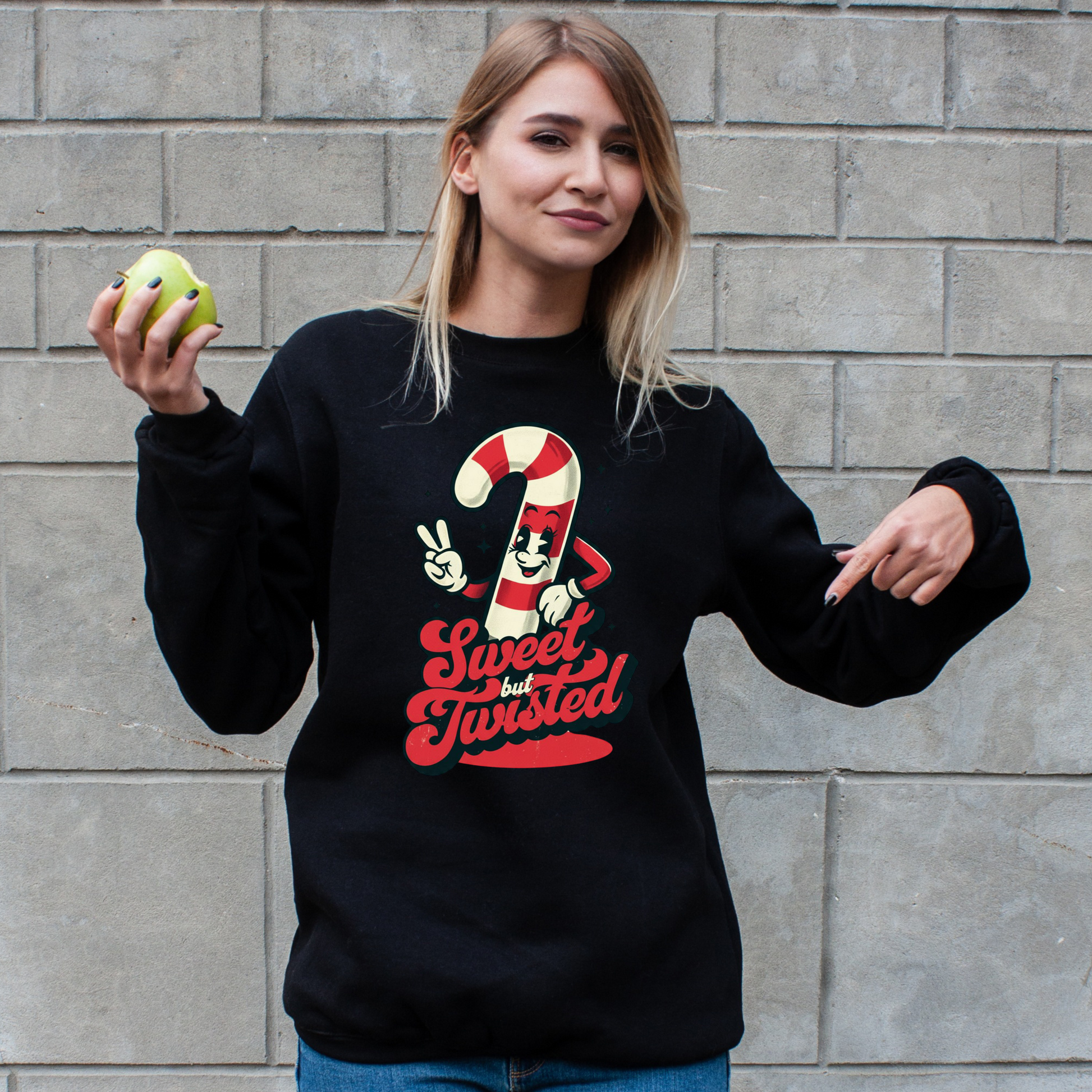 Person wearing a black sweatshirt with a candy cane graphic design and text, holding an apple against a brick wall.