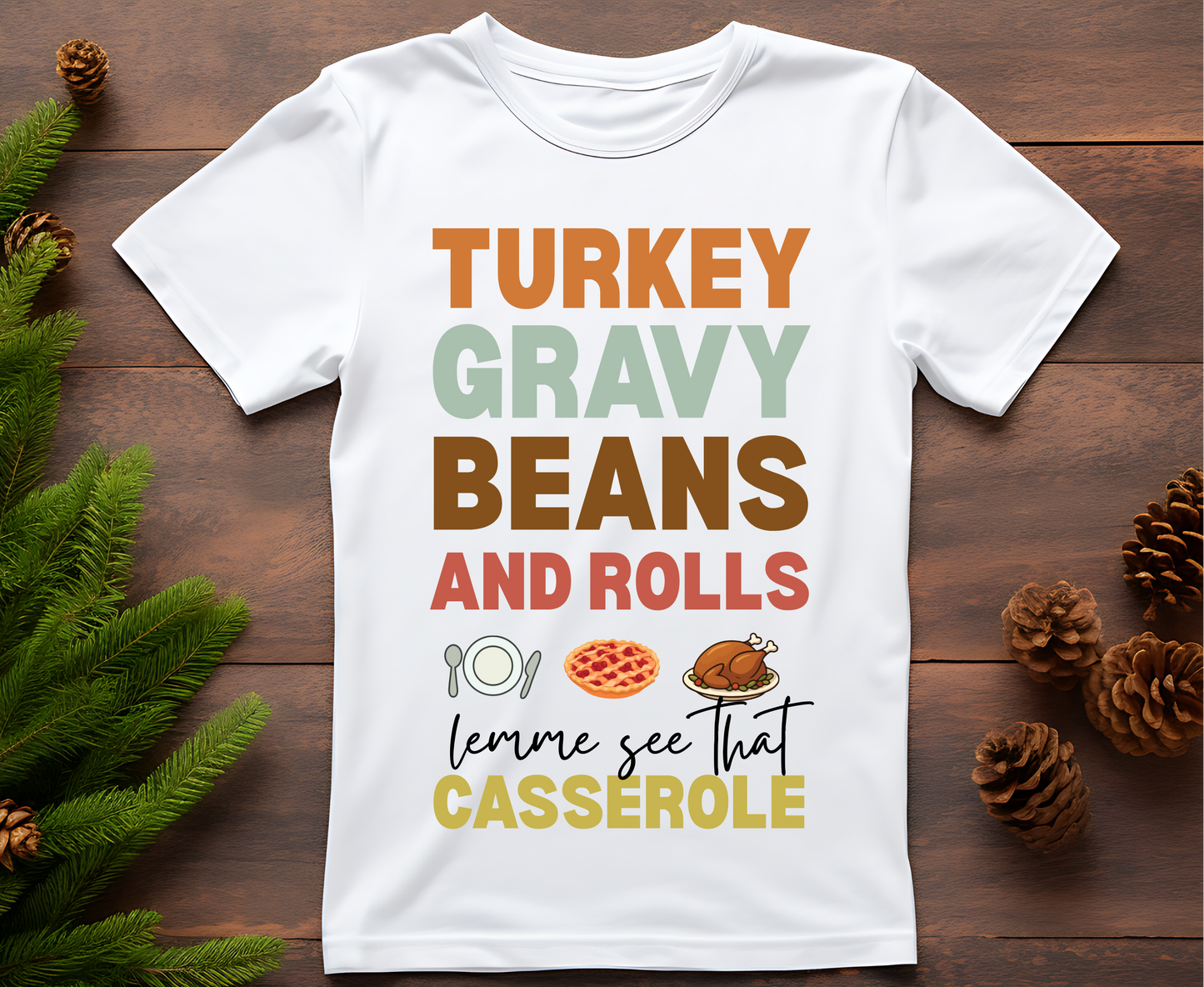 White t-shirt with colorful text and graphics on a wooden surface with pine branches and pinecones.